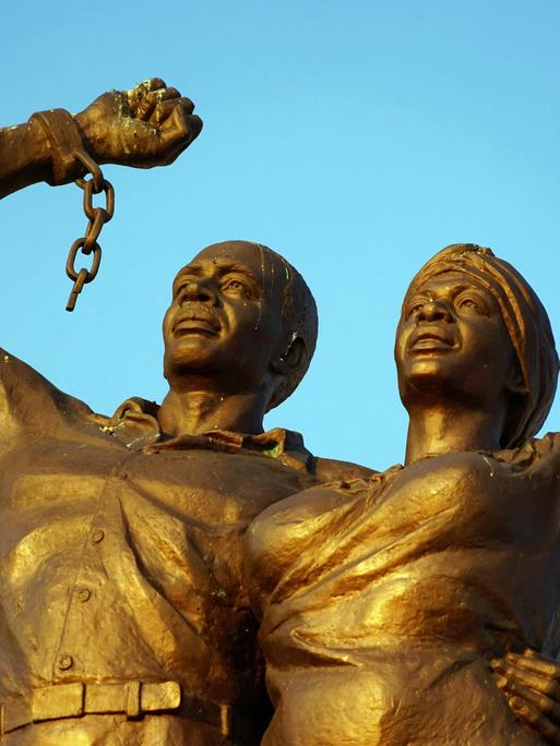 Das riesige Denkmal in Windhoek, Namibia, zeigt einen Mann und eine Frau mit ernstem Gesichtsausdruck, die ihre Arme triumphal gen Himmel strecken. An ihren Handgelenken hängen gesprengte Ketten.