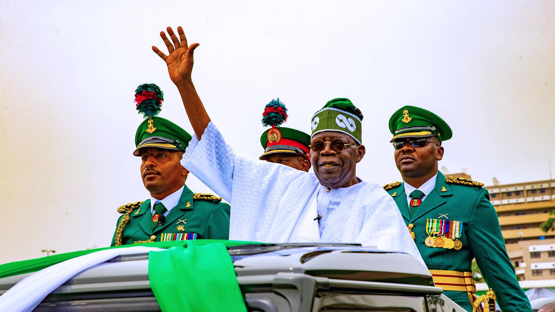 Nigerias Präsident Tinubi winkt aus dem geöffneten Dach eines Autos, hinter stehen mehrere Männer in Uniform.