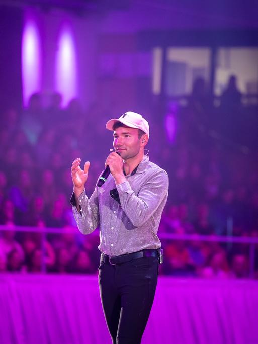 Patrick Thomalla steht in einer Arena und spricht in ein Mikrofon. Er trägt schwarze Reiterhosen und ein Basecap.