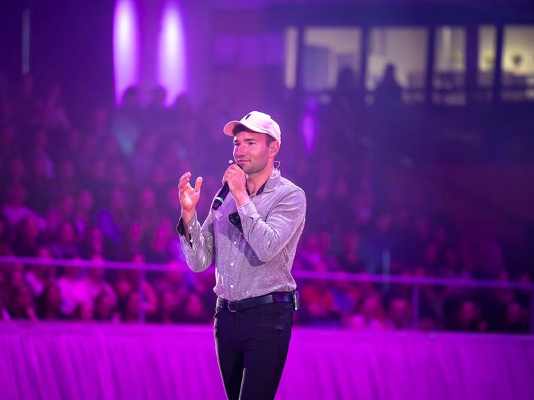 Patrick Thomalla steht in einer Arena und spricht in ein Mikrofon. Er trägt schwarze Reiterhosen und ein Basecap.