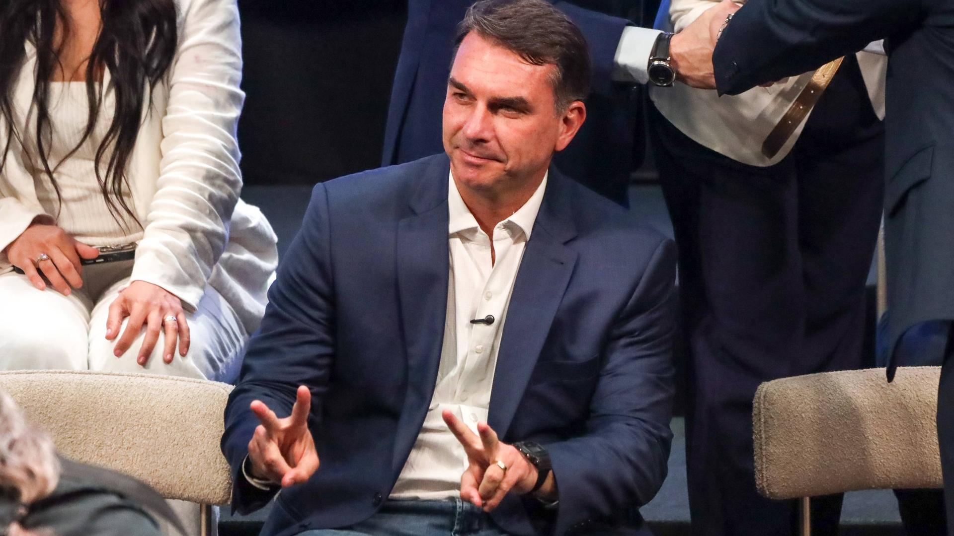Flavio Bolsonaro sitzt in dunkelblauem Blazer, weißen Hemd und Jeans und macht mit beiden Händen das Peace-Zeichen.