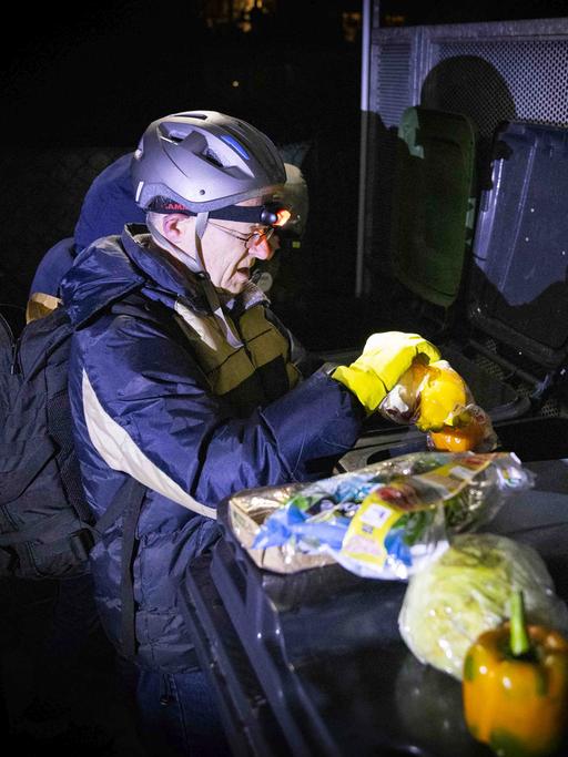 Der Jesuit Jörg Alt ist begleitet mit einer Outdoor-Jacke, er träg auch einen Helm und eine Stirnlampe. Aus einer Mülltonne holt er Lebensmittel.