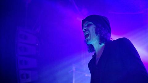 Ein Sänger gibt ein Konzert. Es ist der ehemalige Frontmann der Band HIM: Ville Valo. Er steht auf einer Bühne, der Hintergrund ist in blaues und lila Licht gehüllt.