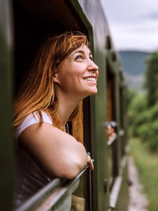 Eine Frau fährt während des Bahnfahrens aus dem Fenster. Der Zug fährt durch eine grüne Landschaft