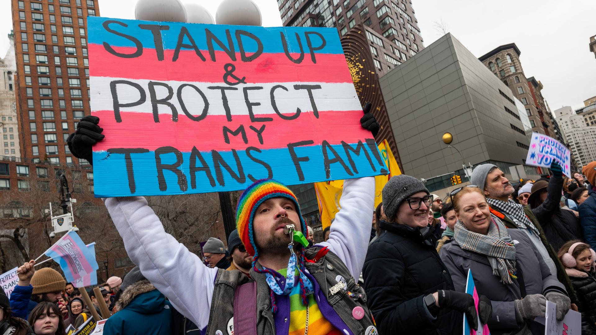 Eine Person mit vielen Regenbogenaccessoires an der Kleidung, einer Sonnenbrille und einem Bart, hält ein Schild hoch, auf dem vor den Farben der Transgender-Pride-Fahne steht „Stand Up & Protect my Trans Fam!“. Eine Person mit vielen Regenbogenaccessoires an der Kleidung, einer Sonnenbrille und einem Bart, hält ein Schild hoch, auf dem vor den Farben der Transgender-Pride-Fahne steht „Stand Up & Protect my Trans Fam!“.