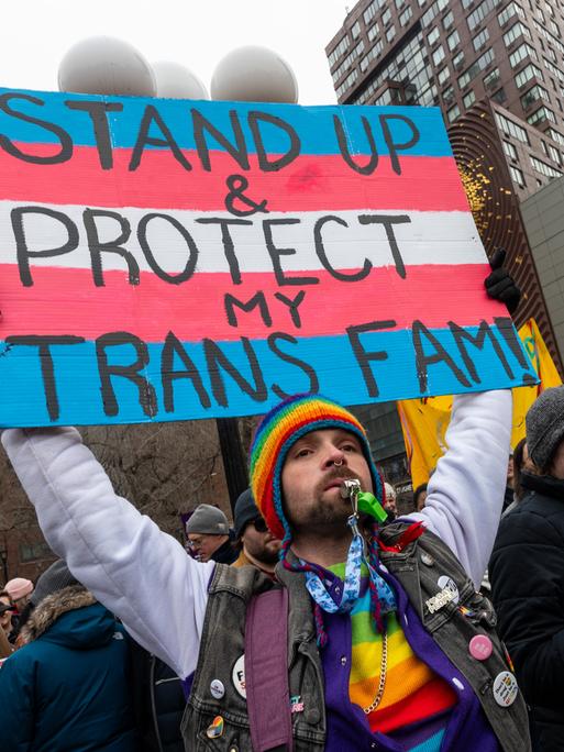 Eine Person mit vielen Regenbogenaccessoires an der Kleidung, einer Sonnenbrille und einem Bart, hält ein Schild hoch, auf dem vor den Farben der Transgender-Pride-Fahne steht „Stand Up & Protect my Trans Fam!“.