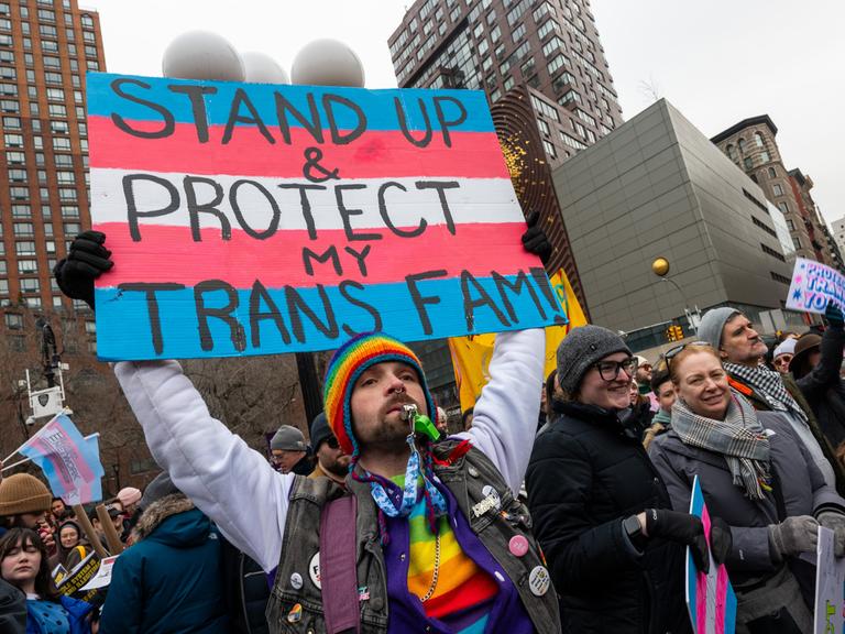 Eine Person mit vielen Regenbogenaccessoires an der Kleidung, einer Sonnenbrille und einem Bart, hält ein Schild hoch, auf dem vor den Farben der Transgender-Pride-Fahne steht „Stand Up & Protect my Trans Fam!“.
