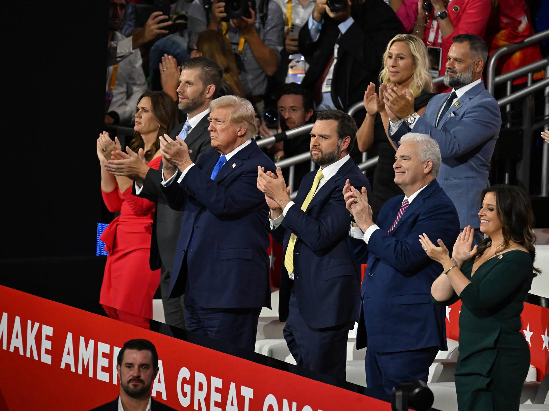 Donald Trump steht mit mehreren Menschen beim Parteitag der Republikaner auf der Tribüne und klatscht. Neben ihm steht J. D. Vance und klatscht ebenfalls. 