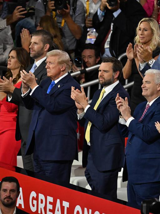 Donald Trump steht mit mehreren Menschen beim Parteitag der Republikaner auf der Tribüne und klatscht. Neben ihm steht J. D. Vance und klatscht ebenfalls. 
