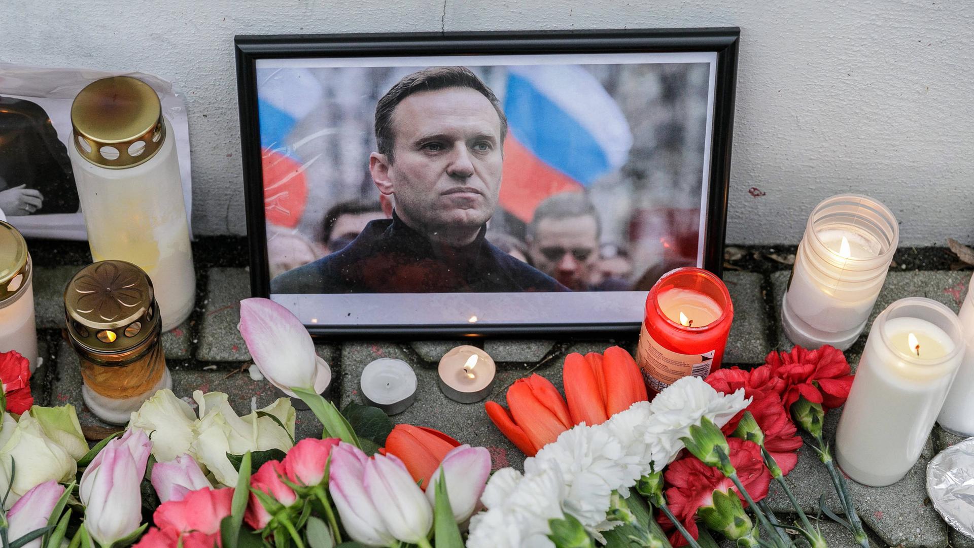 Zwischen Blumen und Kerzen ist das fotografische Porträt eines Mannes zu sehen, im Hintergrund des Fotos die russische Flagge.