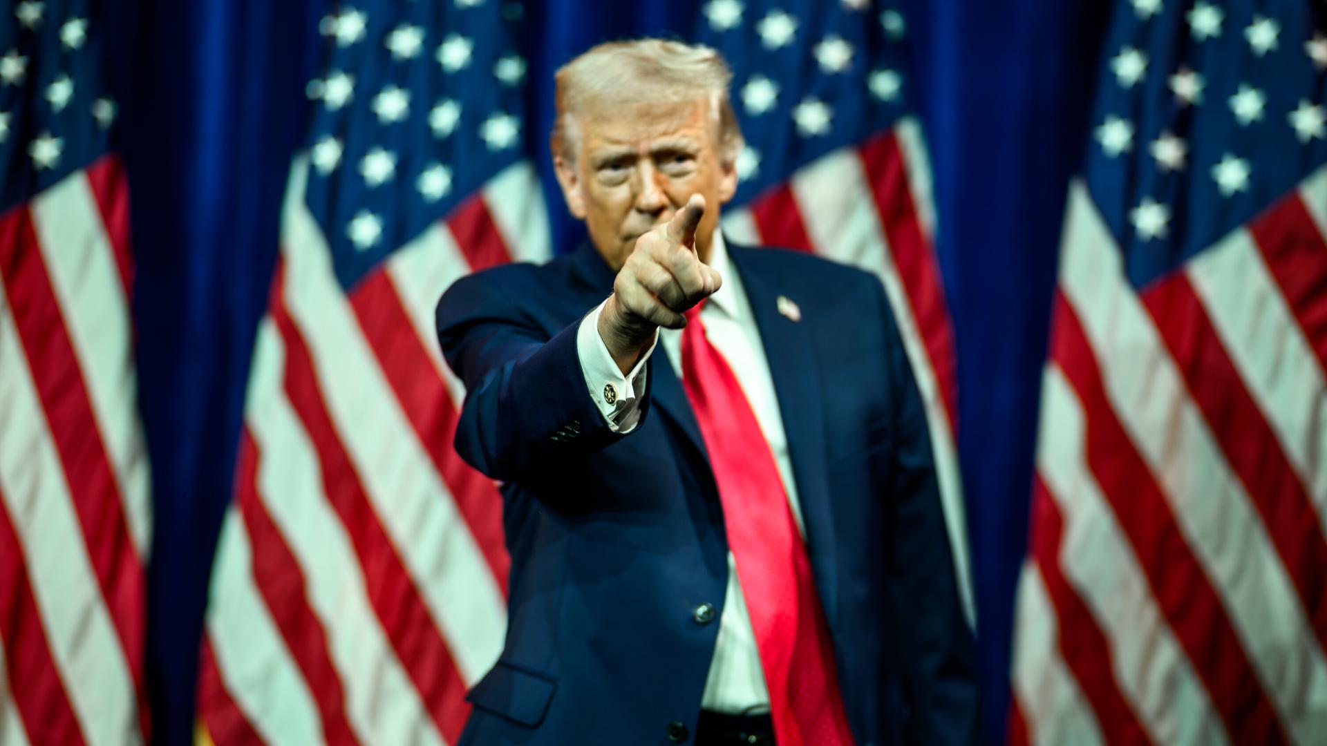 US-Präsident Donald Trump hat den rechten Arm gehoben und deutet mit dem Zeigefinger nach vorne. Im Hintergrund sind mehrere US-Faggen zu sehen.