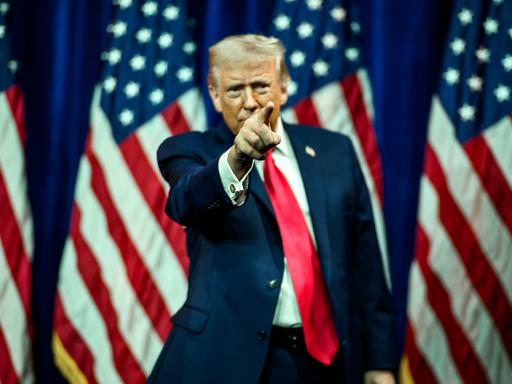 US-Präsident Donald Trump hat den rechten Arm gehoben und deutet mit dem Zeigefinger nach vorne. Im Hintergrund sind mehrere US-Faggen zu sehen.