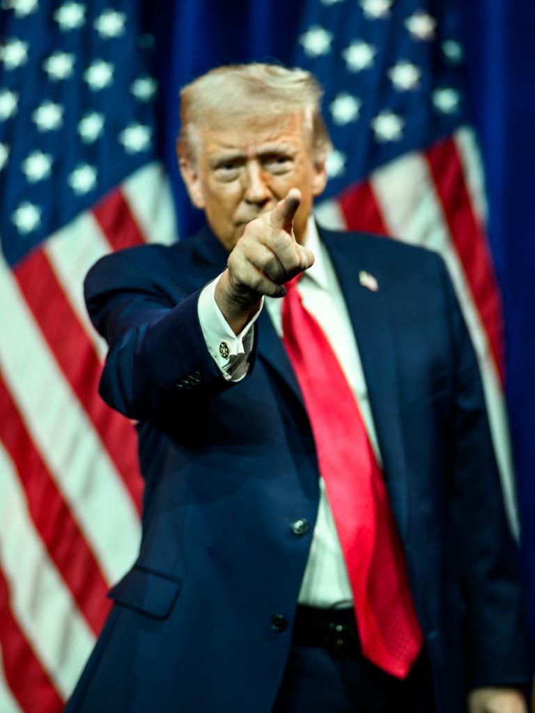 US-Präsident Donald Trump hat den rechten Arm gehoben und deutet mit dem Zeigefinger nach vorne. Im Hintergrund sind mehrere US-Faggen zu sehen.