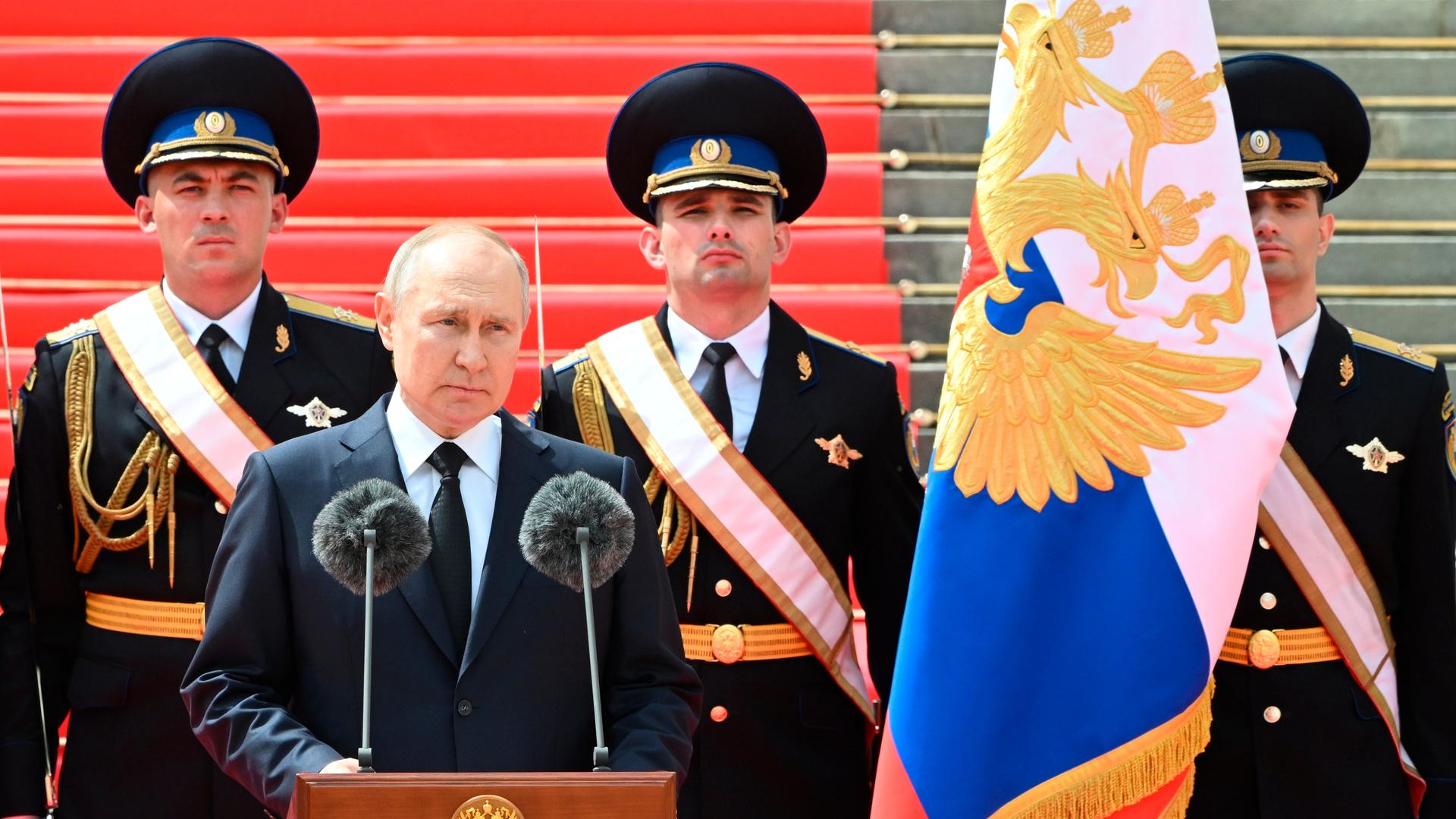Putin spricht an einem Rednerpult. Hinter ihm stehen vor einer Treppe drei Soldaten in schwarzen Paradeuniformen. Rechts neben Putin eine russische Flagge. Putin spricht an einem Rednerpult. Hinter ihm stehen vor einer Treppe drei Soldaten in schwarzen Paradeuniformen. Rechts neben Putin eine russische Flagge.