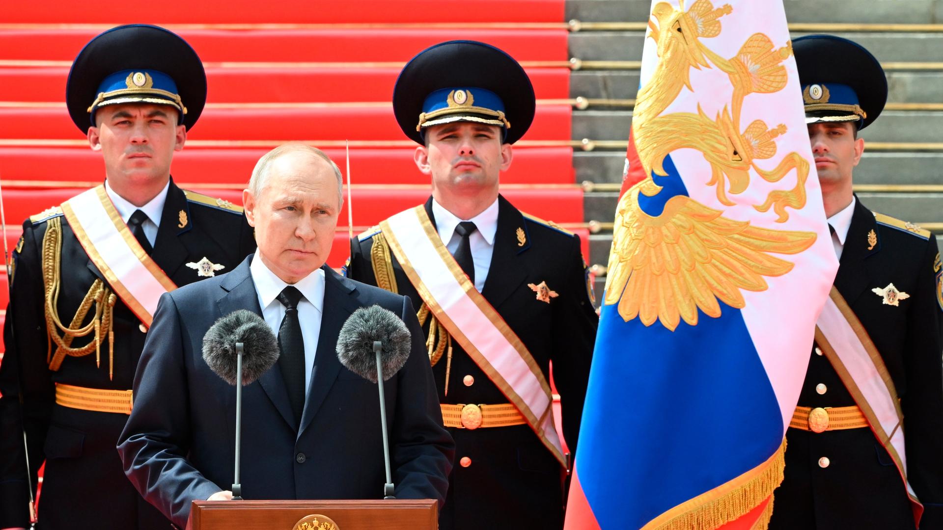 Putin spricht an einem Rednerpult. Hinter ihm stehen vor einer Treppe drei Soldaten in schwarzen Paradeuniformen. Rechts neben Putin eine russische Flagge. 