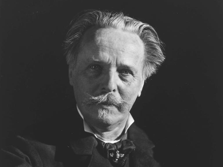 Historisches schwarzweiß Portrait von Karl May der einen geschwungenen Schnurrbart trägt und freundlich in die Kamera schaut. Er sitzt an einem Schreibtisch, seine Hand ruht auf einem Buch.