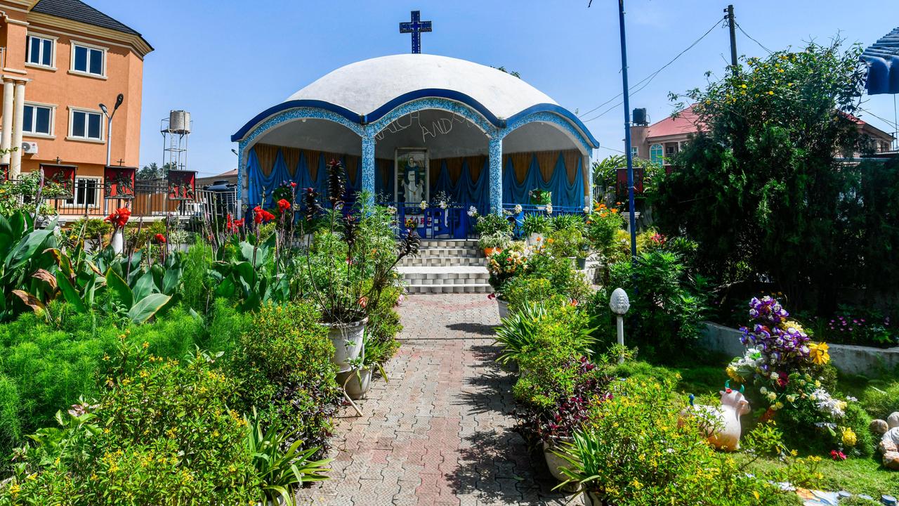 Zu sehen ist eine kleine, zu den Seiten offene Kapelle in runder Bauweise. Auf dem gewölbten Dach ist ein blaues Kruzifix angebracht. Der Vordergrund zeigt einen kurzen Fußweg. Links und rechts davon sind grüne Pflanzen und bunte Blüten zu sehen.