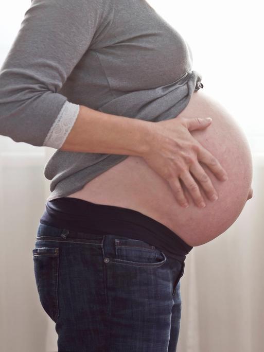 Eine schwangere Frau hält sich den nackten Bauch, der unter einem grauen Oberteil herausguckt