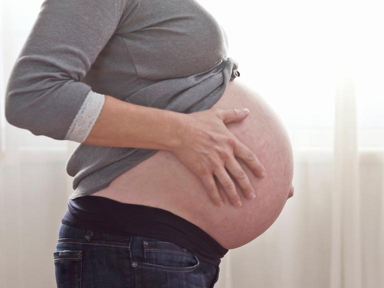 Eine schwangere Frau hält sich den nackten Bauch, der unter einem grauen Oberteil herausguckt