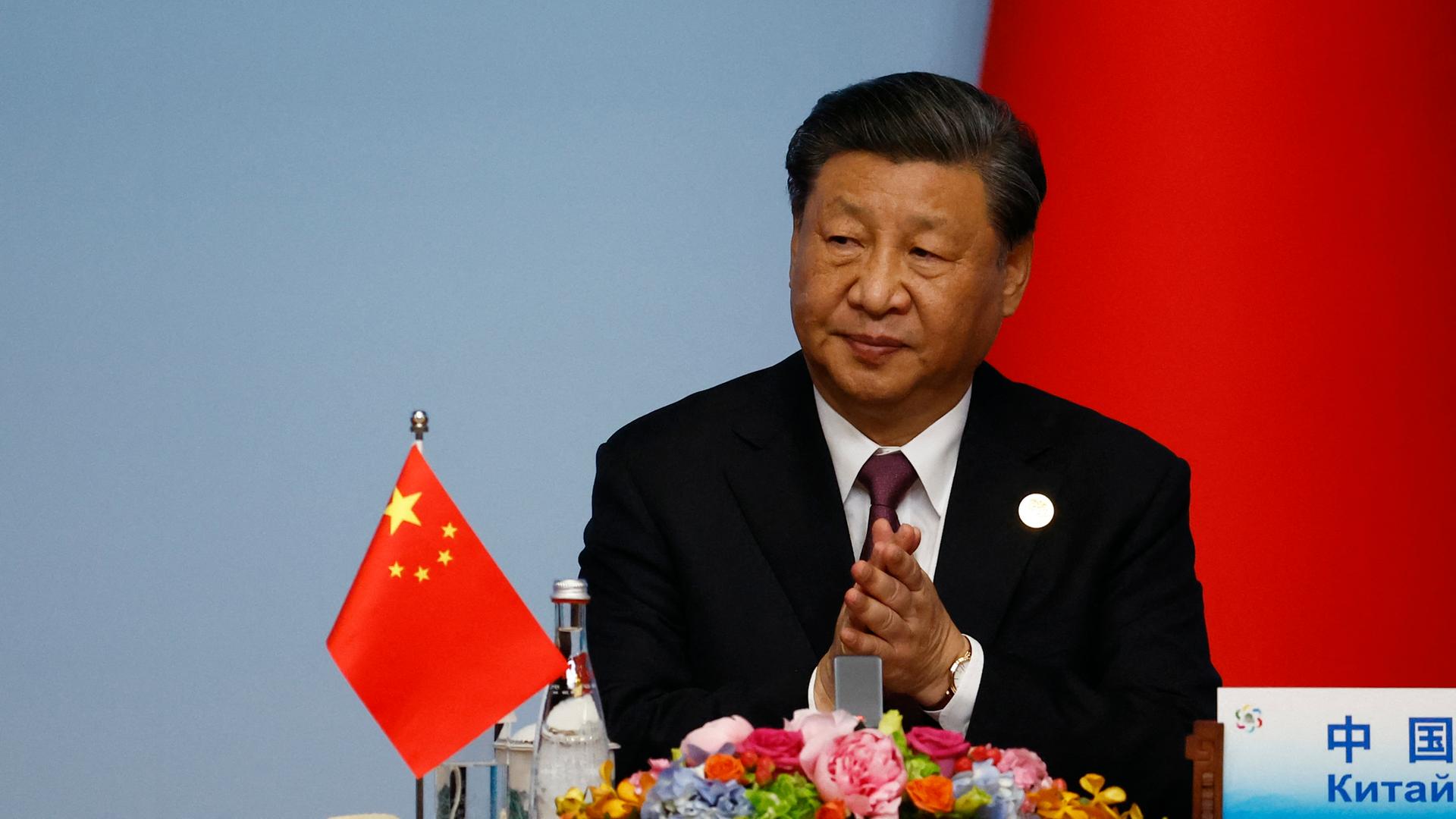 Der chinesische Präsident Xi Jinping sitzt an einem Tisch hinter einem Mikrofon. Auf dem Tisch steht eine chinesische Flagge.