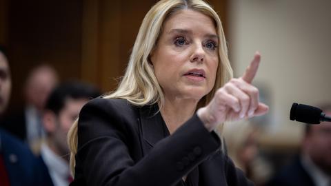 Pam Bondi vor einem Mikrofon. Sie hat den Zeigefinger gehoben.