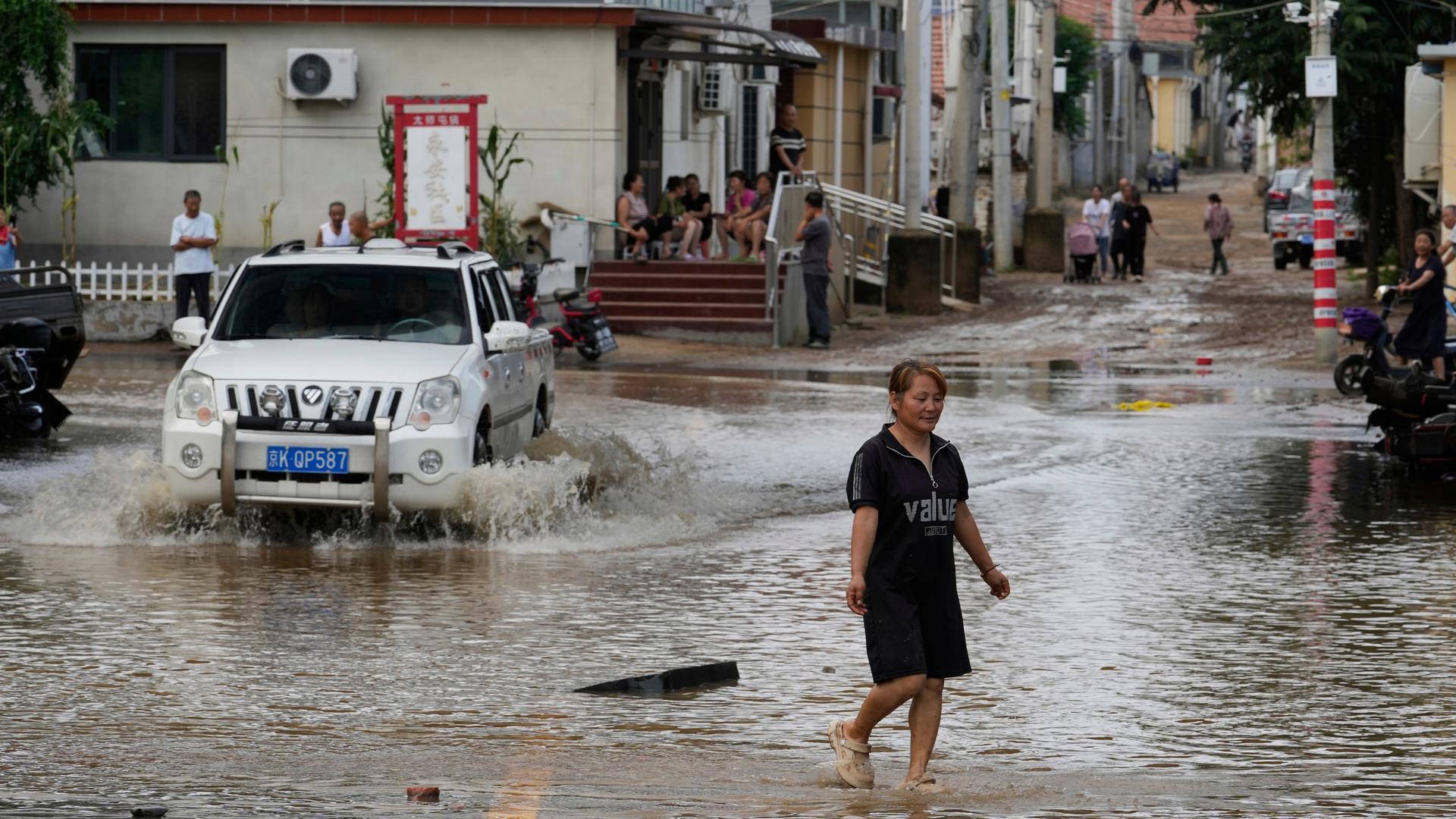 Eine Frau geht durch eine überflutete Straße. Neben ihr fährt ein Auto durch die Wassermassen.