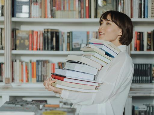Frau trägt einen Stapel Bücher; im Hintergrund steht ein gut gefülltes Bücherregal.
