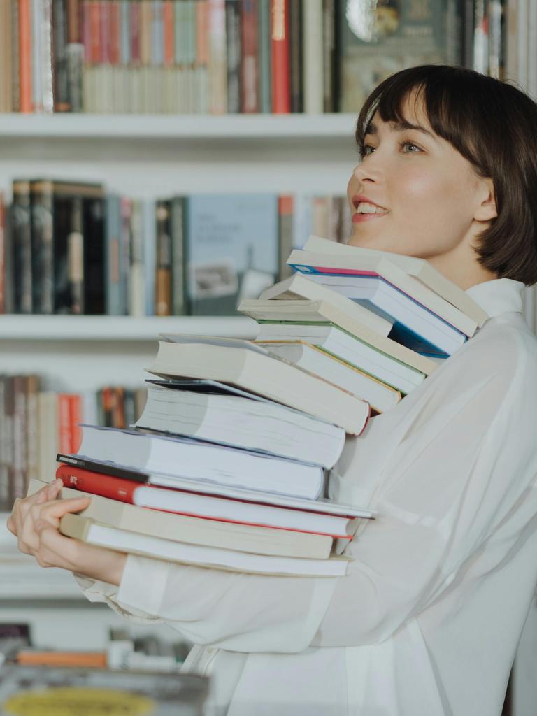 Frau trägt einen Stapel Bücher; im Hintergrund steht ein gut gefülltes Bücherregal.