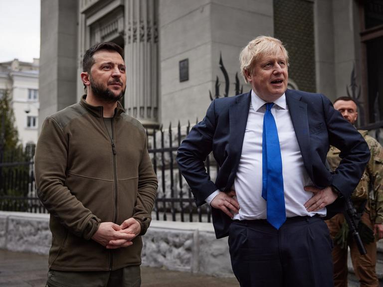 Der damalige britische Prime Minister Boris Johnson und der ukrainische Präsident Wolodymyr Selenskyi gehen über den Unabhängigkeitsplatz in Kiev. Der dunkelhaarige Selenskyi (links) hat einen olivgrünen Pullover an, der blonde Johnson trägt Anzug, weißes Hemd und Krawatte, die Hände stemmt er in die Hüfte.