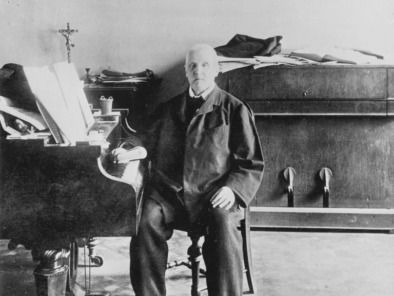 Historisches Schwarzweißporträt von Anton Bruckner, der im dunklen Anzug an einem Klavier sitzt.