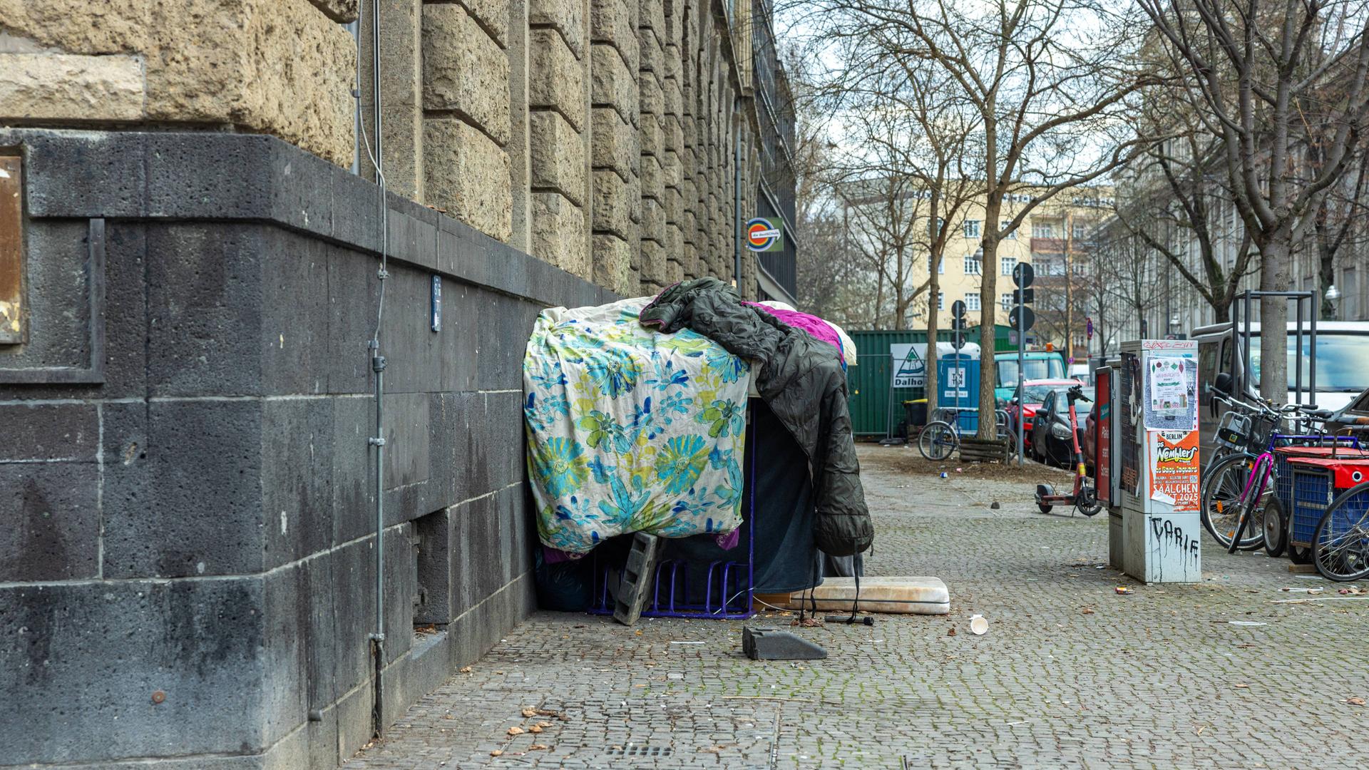 Provisorisch aufgetürmte, schmutzige Bettdecken und Schlafsäcke, die als Unterschlupf für einen Obdachlosen an der Ecke der Ganghoferstraße dienen
