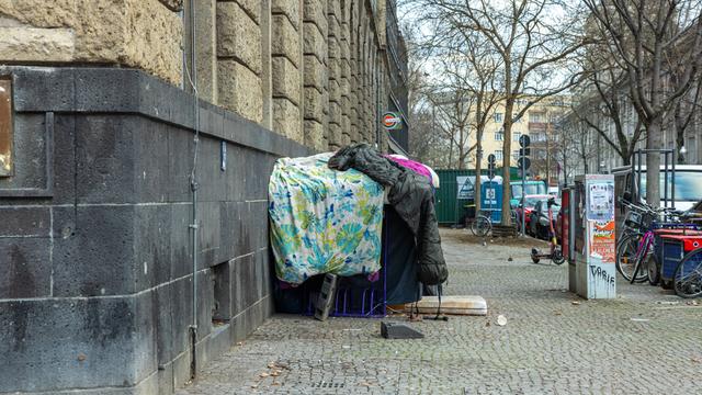 Provisorisch aufgetürmte, schmutzige Bettdecken und Schlafsäcke, die als Unterschlupf für einen Obdachlosen an der Ecke der Ganghoferstraße dienen