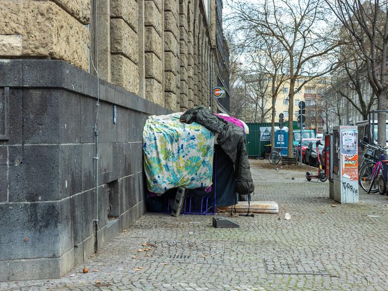 Provisorisch aufgetürmte, schmutzige Bettdecken und Schlafsäcke, die als Unterschlupf für einen Obdachlosen an der Ecke der Ganghoferstraße dienen