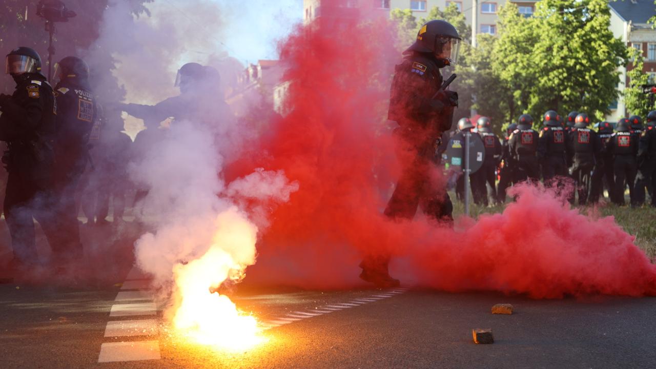In Leipzig laufen Polizisten über eine Straße, auf der Pyrotechnik abbrennt, eine rote Rauchwolke ist zu sehen. In Leipzig laufen Polizisten über eine Straße, auf der Pyrotechnik abbrennt, eine rote Rauchwolke ist zu sehen.