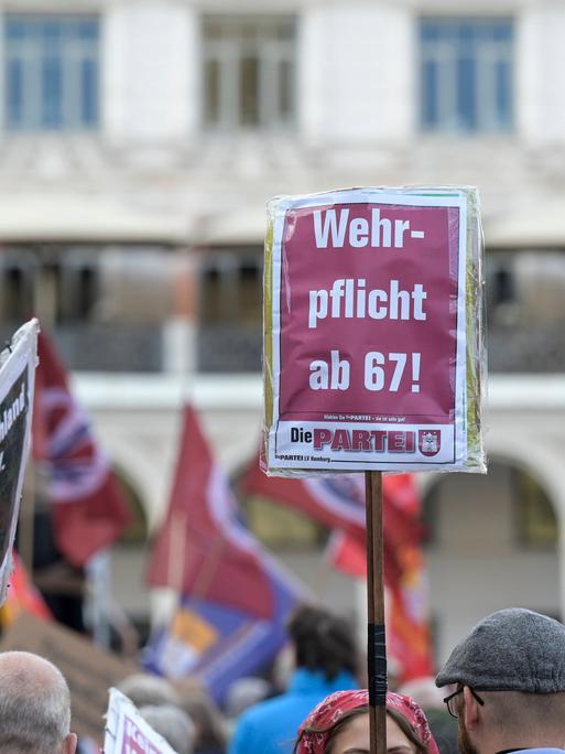 Bei einer Demonstration wird ein Schild von der Partei "Die PARTEI" in die Höhe gehalten. Darauf steht: "Wehrpflicht mit 67!"