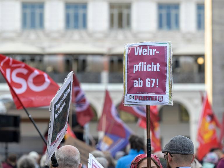 Bei einer Demonstration wird ein Schild von der Partei "Die PARTEI" in die Höhe gehalten. Darauf steht: "Wehrpflicht mit 67!"