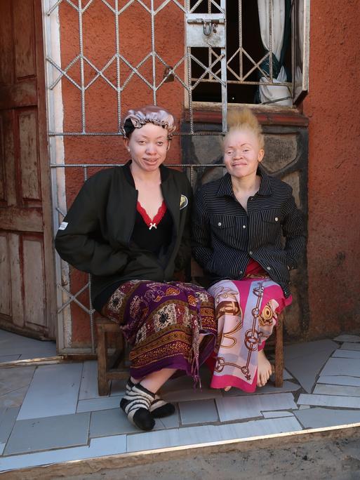 Zwei junge afrikanische Frauen mit für Albinismus charakteristisch heller Hautfarbe sitzen vor einem Haus und lachen in die Kamera.