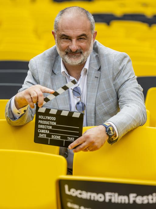 Der künstlerische Leiter des Filmfestivals in Locarno, Giona A. Nazzaro, inmitten von gelben Stühlen mit einer Filmklappe in der Hand. 
