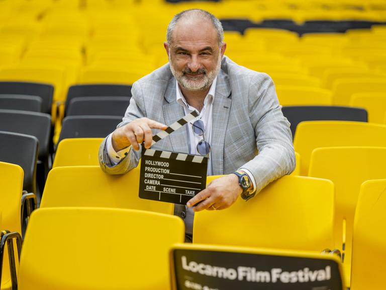 Der künstlerische Leiter des Filmfestivals in Locarno, Giona A. Nazzaro, inmitten von gelben Stühlen mit einer Filmklappe in der Hand. 