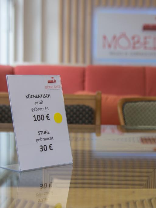 Ein Preisschild auf einem Tisch mit Glasplatte. Auf dem Schild steht: Küchentisch groß, gebraucht, 100 Euro, Stuhl, gebraucht, 30 Euro. Im Hintergrund ist unscharf die Aufschrift "Möbelshop" zu erkennen.
