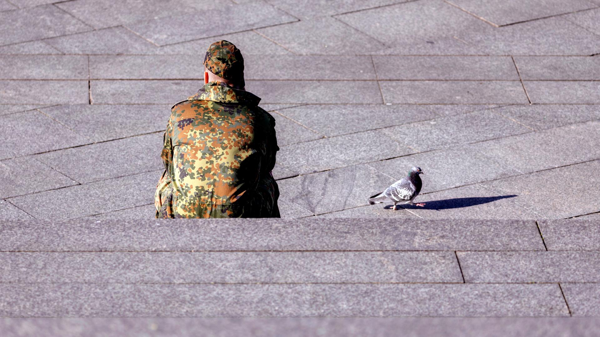 Ein Bundeswehr-Soldat sitzt dem Betrachter mit dem Rücken zugewandt auf einer Treppe. Neben ihm auf dem Beton läuft eine Taube. Ein Bundeswehr-Soldat sitzt dem Betrachter mit dem Rücken zugewandt auf einer Treppe. Neben ihm auf dem Beton läuft eine Taube.