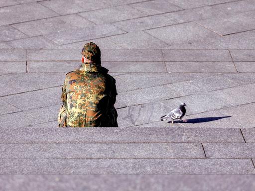 Ein Bundeswehr-Soldat sitzt dem Betrachter mit dem Rücken zugewandt auf einer Treppe. Neben ihm auf dem Beton läuft eine Taube.