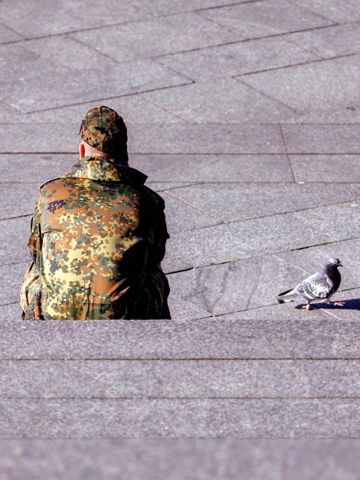Ein Bundeswehr-Soldat sitzt dem Betrachter mit dem Rücken zugewandt auf einer Treppe. Neben ihm auf dem Beton läuft eine Taube.