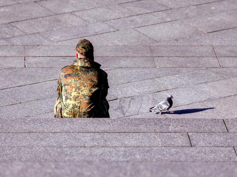 Ein Bundeswehr-Soldat sitzt dem Betrachter mit dem Rücken zugewandt auf einer Treppe. Neben ihm auf dem Beton läuft eine Taube.