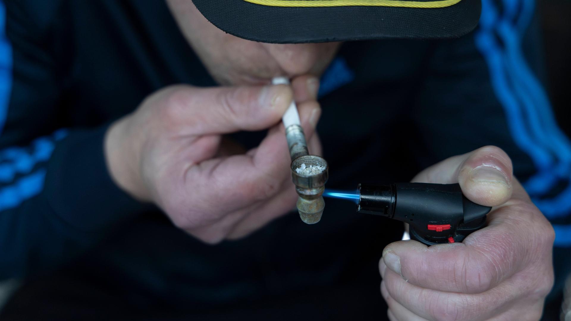 Ein junger Mann raucht Crack und hält dazu eine kleine Metallpfeife in der Hand. Mit der anderen hält er einen kleinen Gasbrenner, um das Crack zu erhitzen.
