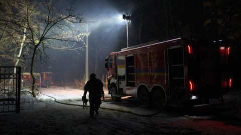 Es ist dunkel. Ein Feuerwehrauto beleuchtet ein Gelände neben einem Gebäude; auf dem Boden liegt ein Löschschlauch. Im Vordergrund die Silhouette eines Feuerwehrmanns. 