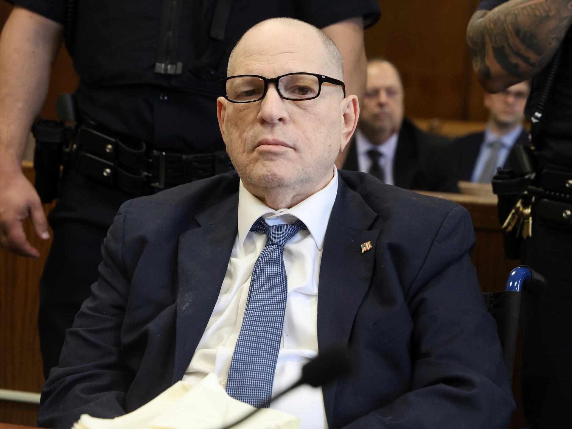 Ein älterer Mann mit Brille sitzt in einem Gerichtsaal und blickt ernst in die Kamera.