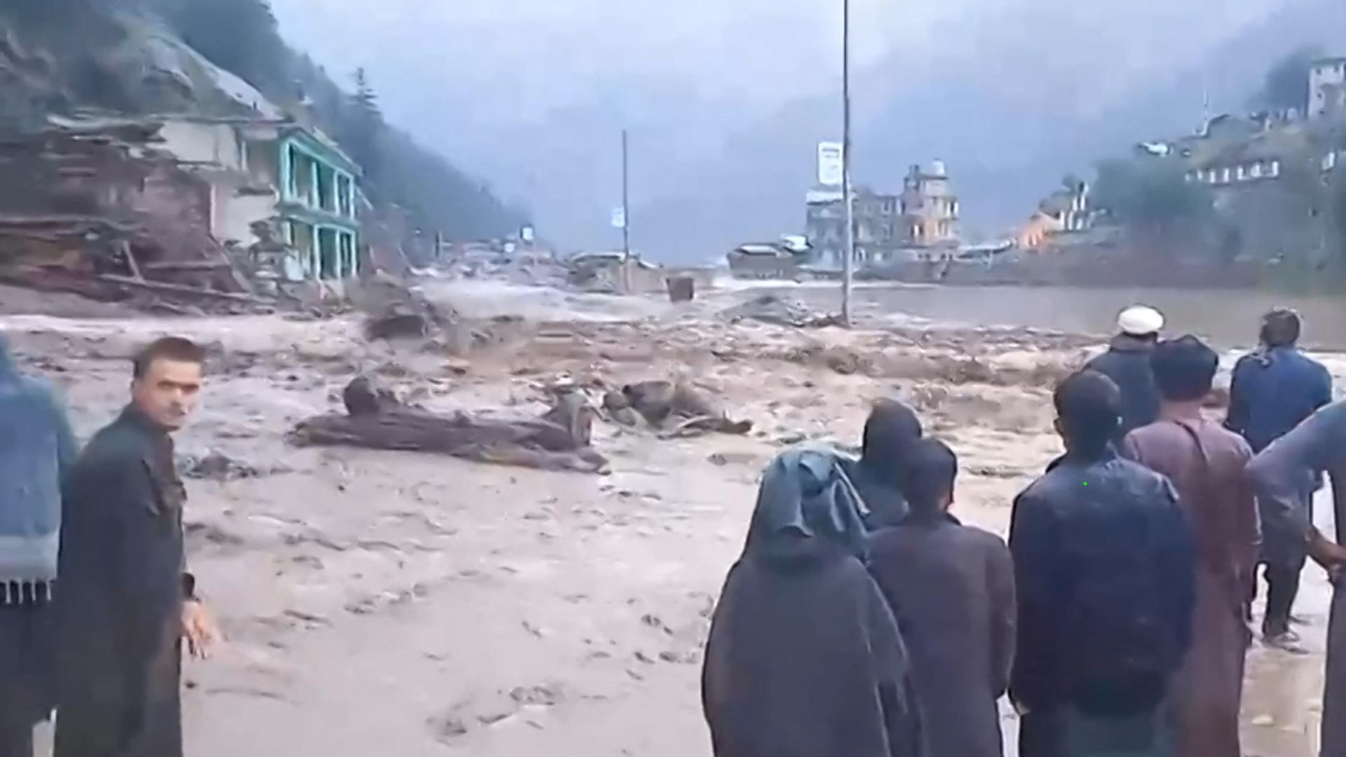 Das Bild zeigt mehrere Personen, die auf Überflutungen schauen. Auf einer Straße steht viel Wasser. Das Bild zeigt mehrere Personen, die auf Überflutungen schauen. Auf einer Straße steht viel Wasser.
