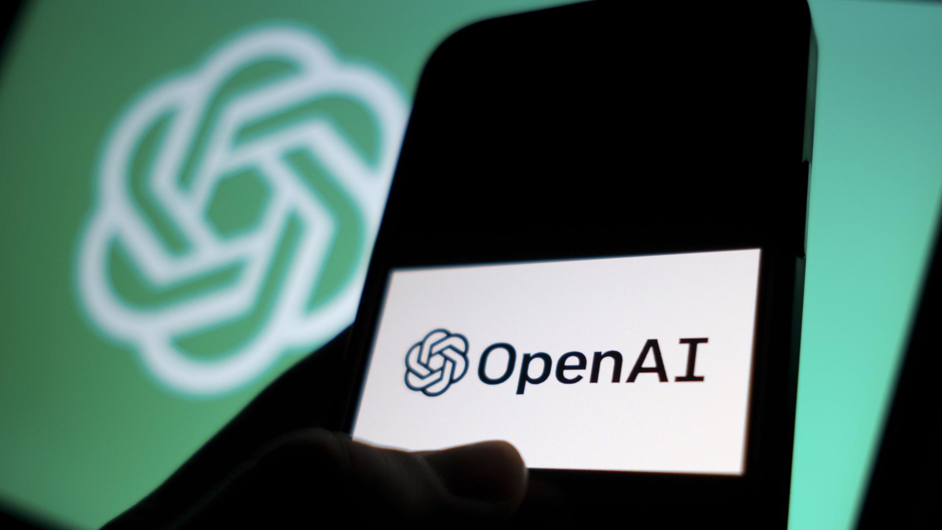 DAs Logo von"OpenAI" auf einem Smartphone, dahinter das Logo der Texterzeugungs-Ki auf einem Computer-Bildschirm.