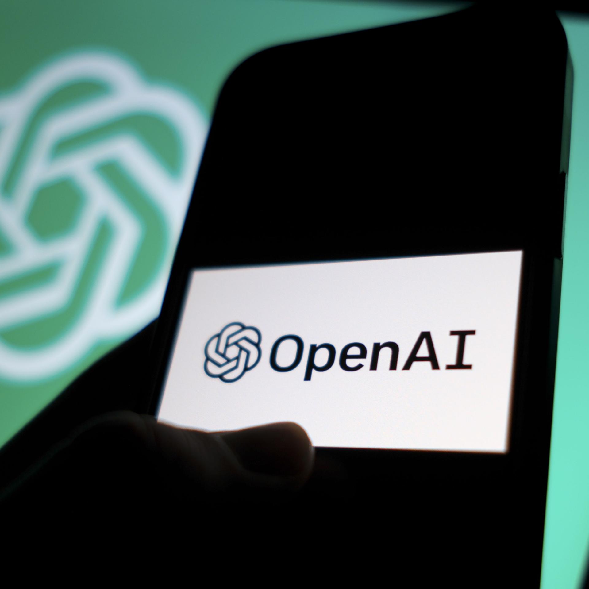 DAs Logo von"OpenAI" auf einem Smartphone, dahinter das Logo der Texterzeugungs-Ki auf einem Computer-Bildschirm